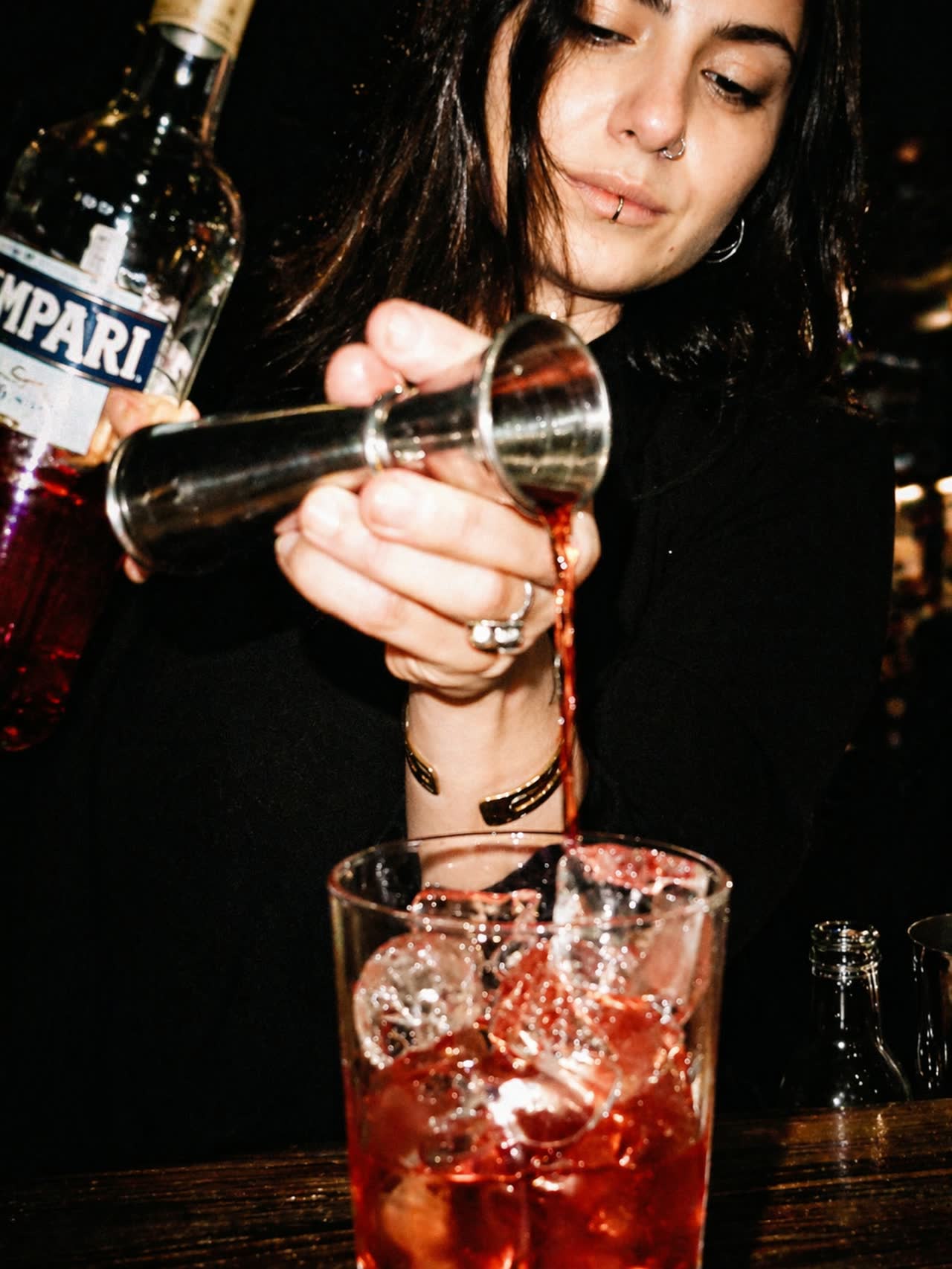 Una bartender Night Wave versa dal jigger in un cocktail al banco, con luce diretta e fondo scuro.